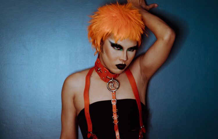 Aggressive young ethnic androgynous man in BDSM accessories standing near blue wall
